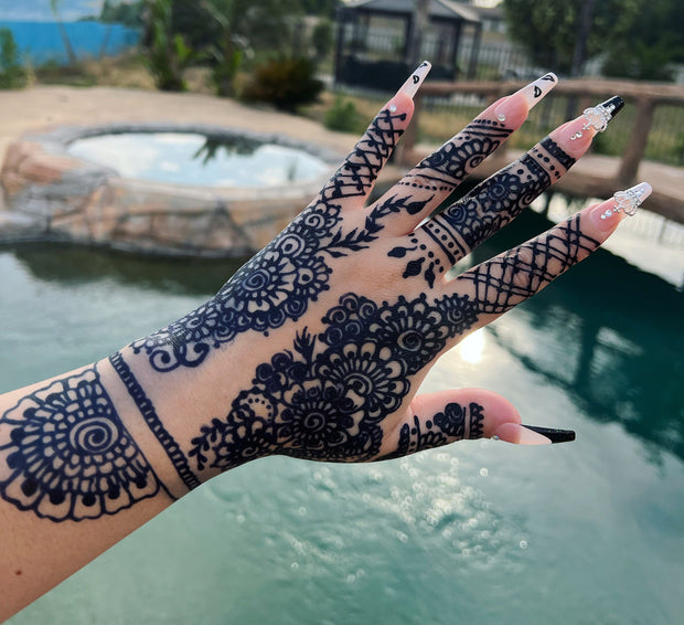 A woman's hand with an intricate jagua design extending from the hand to the wrist, featuring a deep blue-black stain on fair skin. She has acrylic nails. The design was created using Amarya Henna’s natural jagua cones