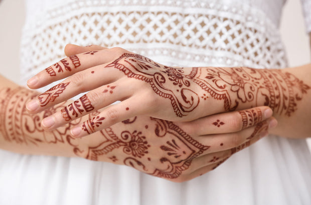 A woman's hands with a deep red-stained henna design, created using Amarya Henna cones. Her hands are crossed in front of a white shirt