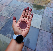 A woman's palm and fingertips adorned with a traditional African-style henna design, featuring a large circle in the center of the palm and dots leading up the fingers, with deep red-stained fingertips. She is wearing a watch that highlights a bold circular face on her wrist.
