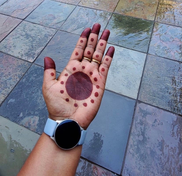 A woman's palm and fingertips adorned with a traditional African-style henna design, featuring a large circle in the center of the palm and dots leading up the fingers, with deep red-stained fingertips. She is wearing a watch that highlights a bold circular face on her wrist.