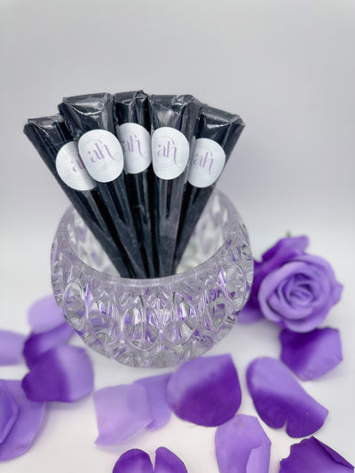 A collection of jagua henna cones labeled 'Amarya Henna' arranged in a clear glass dish with purple rose petals scattered around.