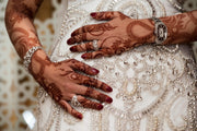 Bride in white wedding dress with hands featuring intricate deep red henna designs, adorned with rings, bracelets, and other jewelry, resting on her gown. Cones from Amarya Henna