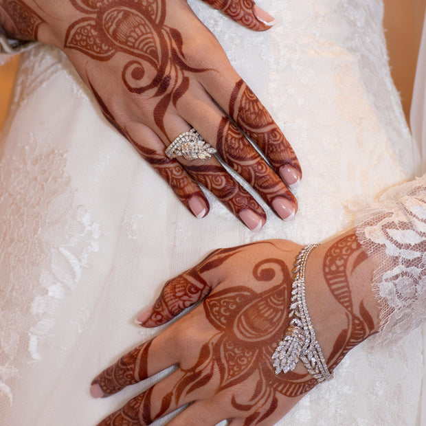Bride in white wedding dress with hands featuring intricate deep red henna designs, adorned with rings, bracelets, and other jewelry, resting on her gown.