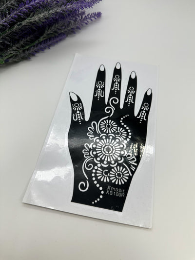 A black and white image of a hand with a henna stencil design on it, placed on a white surface, with a purple flower in the background.