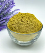A clear glass bowl filled with Pure Rajasthani Amarya Henna powder, placed on a white surface with a lavender flower arrangement in the background.