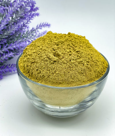 A clear glass bowl filled with Pure Rajasthani Amarya Henna powder, placed on a white surface with a lavender flower arrangement in the background.